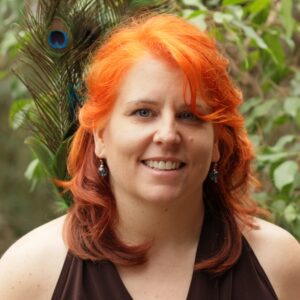 Portrait of Shanya Luther, a woman with red hair, wearing a black top and earrings, in front of foliage and a peacock feather.