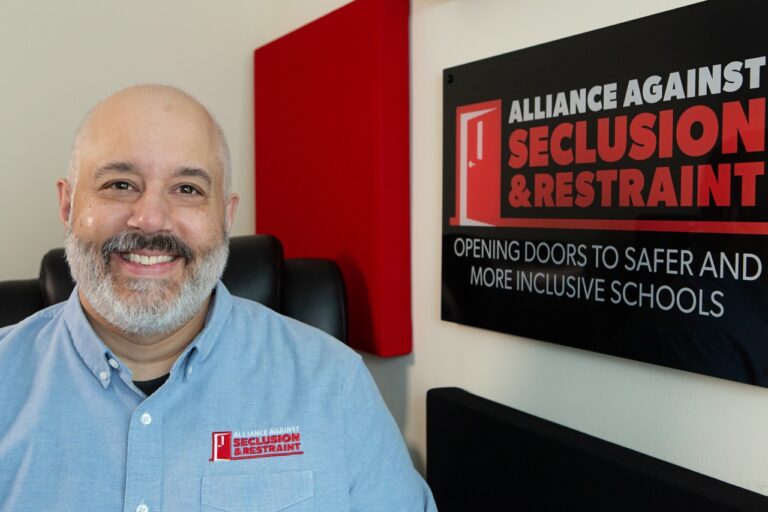 Guy Stephens, a middle-aged white man with bald head, dark mustache and close cropped white beard smiles at the camera beside a wall sign that says Alliance Against Seclusion & Restraint, Opening doors to safer and more inclusive schools